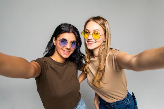 Portrait of two lovely women in sunglasses standing and taking a selfie isolated over white wall