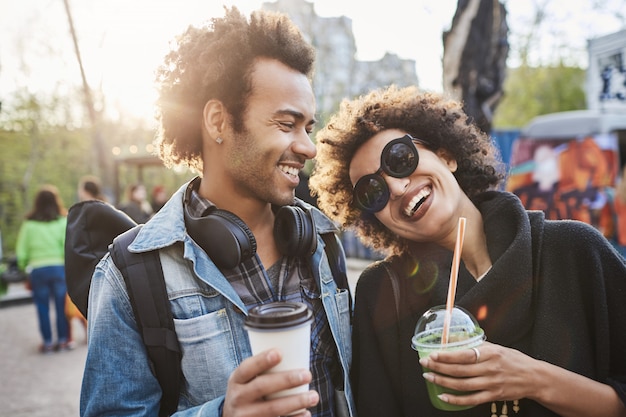 Portrait of two lovers with afro haircuts, strolling in park and drinking coffee while talking and enjoying spending time at food festival.