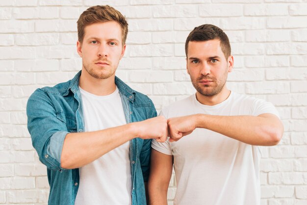 Portrait of a two male friends bumping fist standing against white brick wall