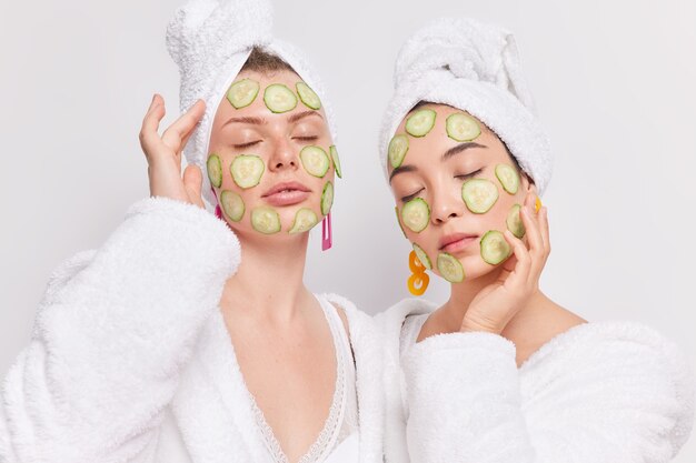 Portrait of two mixed race women stand with closed eyes apply cucumber slices on face to moisturize enjoy softness of skin 