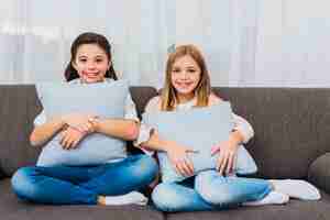 Free photo portrait of two smiling girls sitting on sofa with blue cushions in hand looking to camera