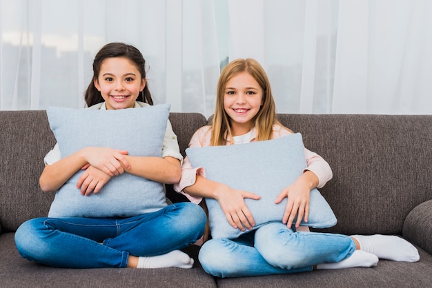 Portrait of two smiling girls sitting on sofa with blue cushions in hand looking to camera