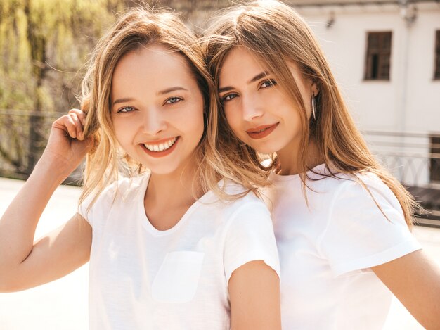 Портрет двух молодых красивых белокурых улыбающихся хипстерских девочек в модной летней белой футболке одевается. , Позитивные модели с удовольствием. Обниматься