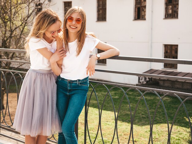 Portrait of two young beautiful blond smiling hipster girls in trendy summer white t-shirt clothes.   . Positive models having fun in sunglasses.Hugging