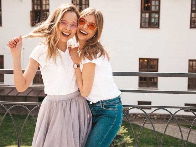 Un ritratto di due giovani belle ragazze sorridenti bionde dei pantaloni a vita bassa in vestiti bianchi della maglietta dell'estate d'avanguardia. . modelli positivi che si divertono in occhiali da sole