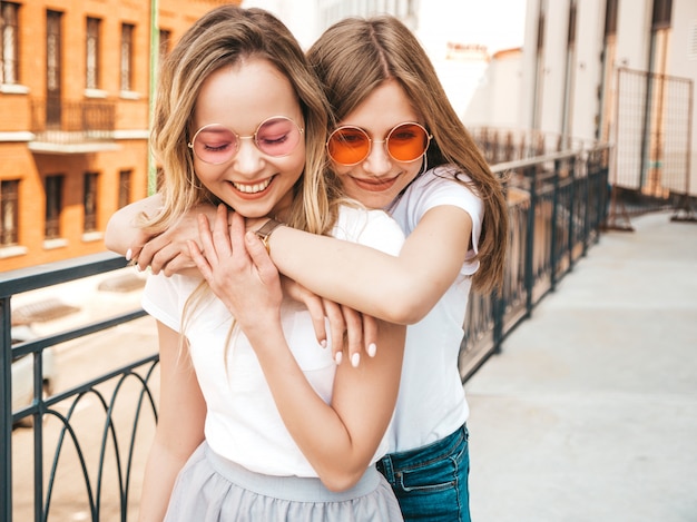 Portrait of two young beautiful blond smiling hipster girls in trendy summer white t-shirt clothes.   . Positive models having fun in sunglasses.Hugging