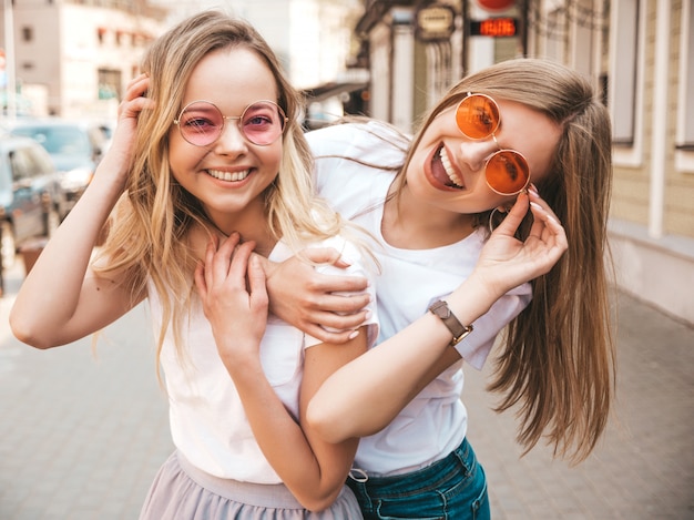 トレンディな夏の白いtシャツ服で流行に敏感な女の子を笑顔2つの若い美しいブロンドの肖像画。 。サングラスを楽しんでいるポジティブなモデル。