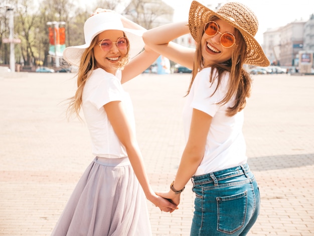 Портрет двух молодых красивых белокурых улыбающихся хипстерских девочек в модной летней белой футболке одевается. , Позитивные модели, держа друг друга за руки
