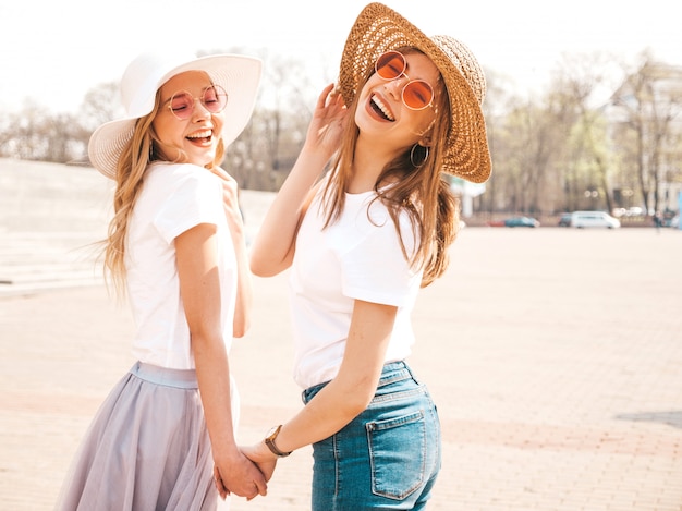 Портрет двух молодых красивых белокурых улыбающихся хипстерских девочек в модной летней белой футболке одевается. , Позитивные модели, держа друг друга за руки