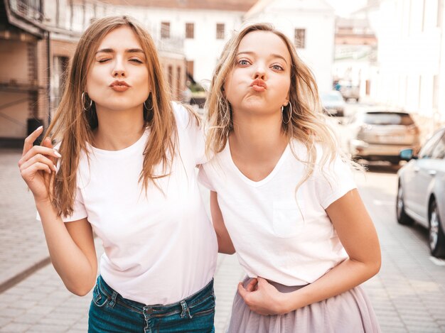 Portrait of two young beautiful blond smiling hipster girls in trendy summer white t-shirt clothes.  . Positive models makes duck face