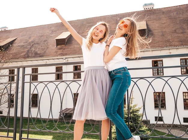 Portrait of two young beautiful blond smiling hipster girls in trendy summer white t-shirt clothes.   . Positive models raising hands