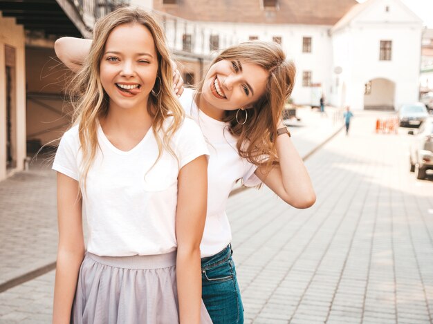 Portrait of two young beautiful blond smiling hipster girls in trendy summer white t-shirt clothes.  . Positive models shows tongue