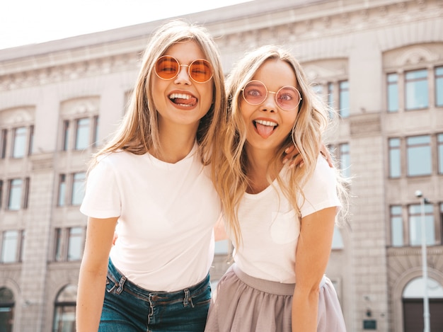 Un ritratto di due giovani belle ragazze sorridenti bionde dei pantaloni a vita bassa in vestiti bianchi della maglietta dell'estate d'avanguardia. donne spensierate sexy in posa sulla strada. modelli positivi che mostrano la lingua, in occhiali da sole