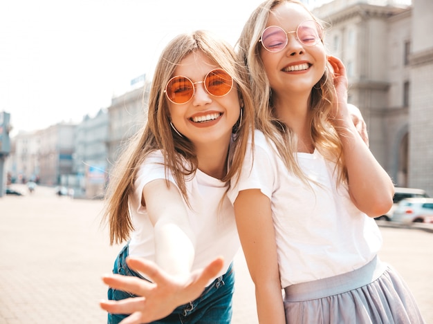 Un ritratto di due giovani belle ragazze sorridenti bionde dei pantaloni a vita bassa in vestiti bianchi della maglietta dell'estate d'avanguardia.