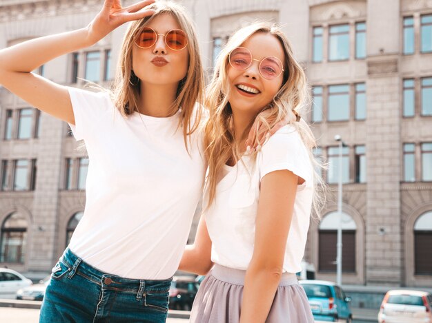 Портрет двух молодых красивых белокурых улыбающихся хипстерских девочек в модной летней белой футболке одевается.
