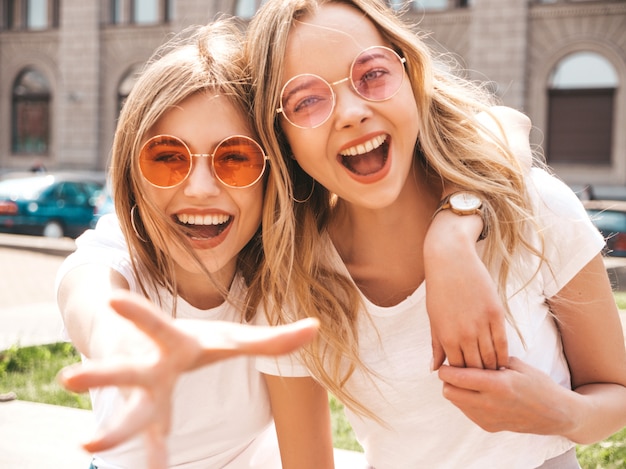 Foto gratuita un ritratto di due giovani belle ragazze sorridenti bionde dei pantaloni a vita bassa in vestiti bianchi della maglietta dell'estate d'avanguardia.