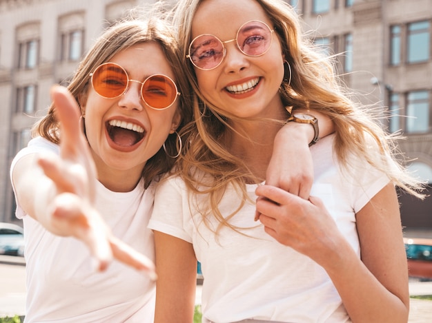 Portrait of two young beautiful blond smiling hipster girls in trendy summer white t-shirt clothes.  