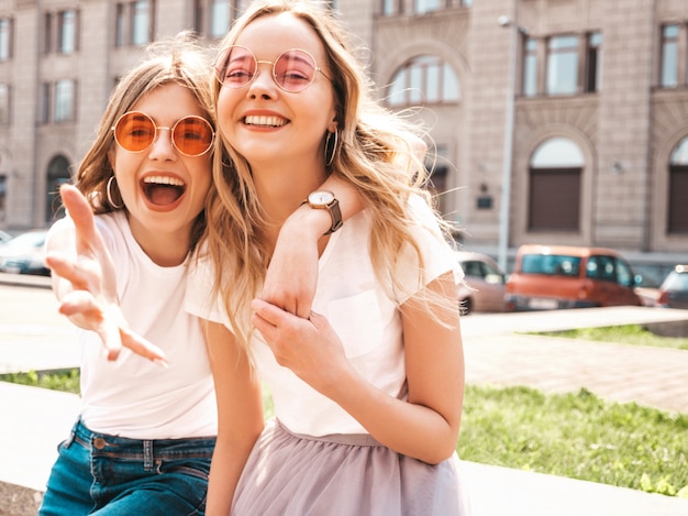 Un ritratto di due giovani belle ragazze sorridenti bionde dei pantaloni a vita bassa in vestiti bianchi della maglietta dell'estate d'avanguardia.