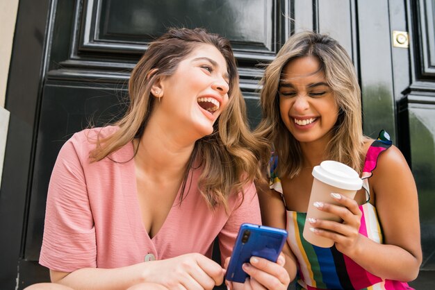 Portrait of two young friends enjoying together and using their mobile phone while sitting outdoors. Urban concept.