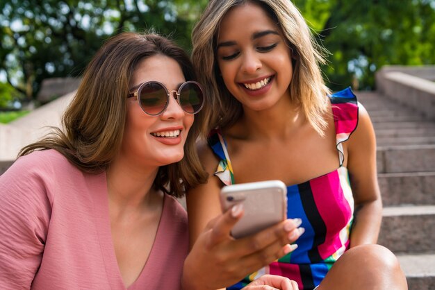 Portrait of two young friends having fun together and using their mobile phone while sitting outdoors