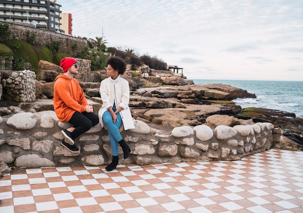 Portrait of two young friends spending good time together and having a conversation while sitting with the sea in the space.