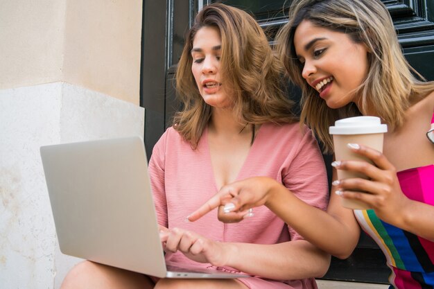 Portrait of two young friends using a laptop and drinking coffee while sitting outdoors. Urban concept. Technology concept.