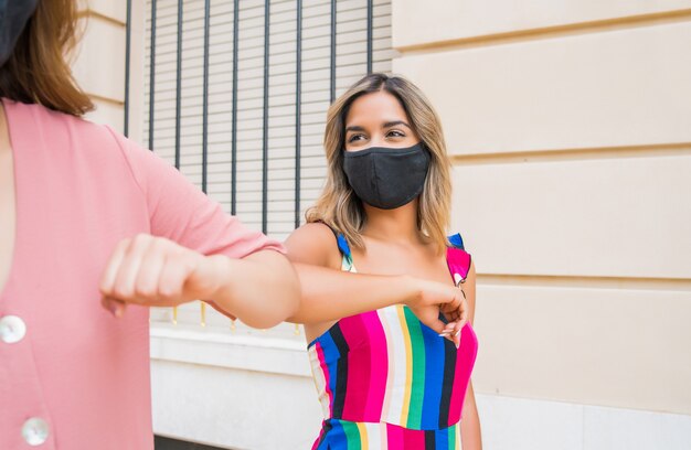 Portrait of two young friends wearing face mask and bumping elbows outdoors. 