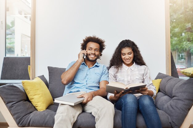 Portrait of two young good-looking dark-skinned students in casual clothes sitting together on sofa in modern library, reading books, preparing for exams, talking o phone.