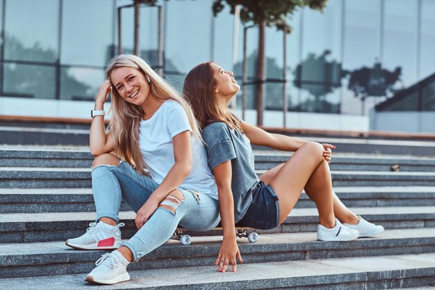 Портрет двух молодых хипстерских девушек, сидящих вместе на скейтборде на ступеньках на фоне небоскреба.