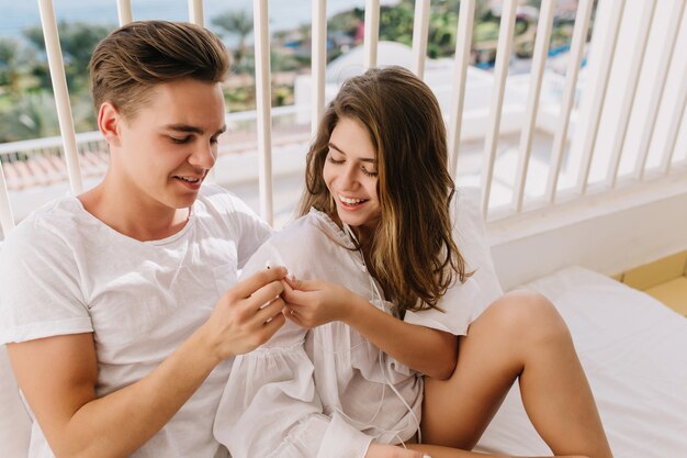 Portrait of two young loving people in white clothes sitting on balcony in sunny morning and listening music in earphones. Cheerful tanned girl gives boyfriend headphone to enjoy cool song together