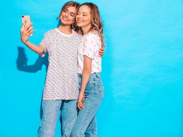 Portrait of two young stylish smiling blond women. Girls dressed in summer hipster clothes. Positive models making selfie on smartphone near blue wall in studio