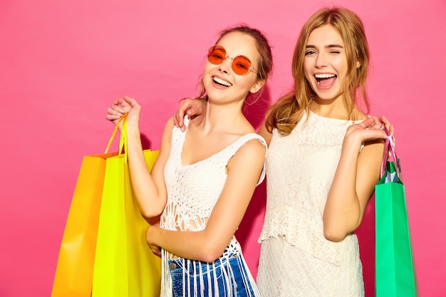 買い物袋を保持している2人の若いスタイリッシュな笑顔金髪女性の肖像画。夏の流行に敏感な服を着た女性。ピンクblackgroundでポーズポジティブモデル