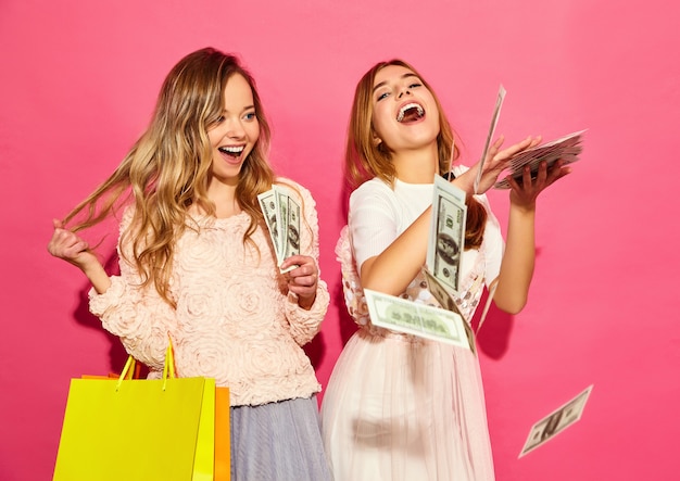 買い物袋を保持している2人の若いスタイリッシュな笑顔金髪女性の肖像画。夏の流行に敏感な服を着た女性。ピンクの壁にお金をかける肯定的なモデル