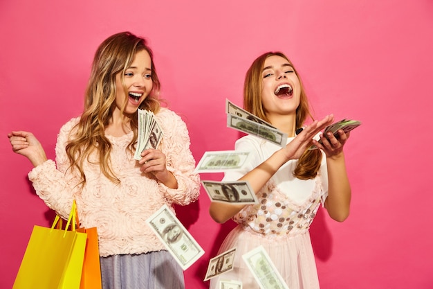 쇼핑백을 들고 두 젊은 세련 된 웃는 금발 여자의 초상화. 여자 여름 hipster 옷을 입고. 분홍색 벽에 돈을 쓰는 긍정적 인 모델