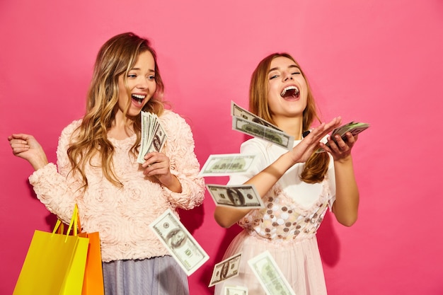 Portrait of two young stylish smiling blond women holding shopping bags. women dressed in summer hipster clothes. Positive models spending money over pink wall