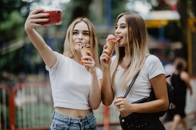 Portrait of two young women standing together eating ice cream and taking selfie in summer street.