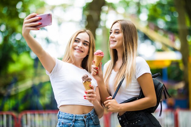 Portrait of two young women standing together eating ice cream and taking selfie in summer street.