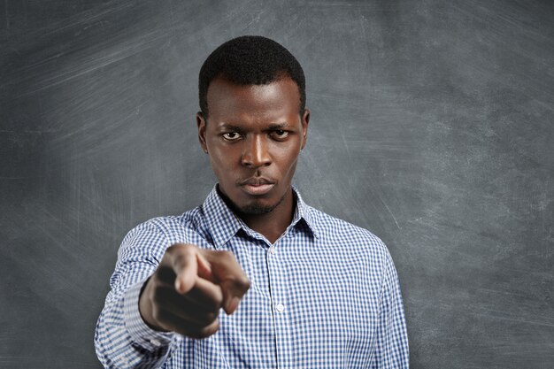 Portrait of unhappy African boss having mad expression pointing his index finger, looking angrily and frowning as if accusing or blaming you for mistake. Selective focus on man's face