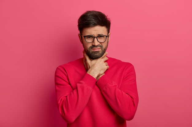 Portrait of unhappy bearded Caucasian man chocks himself, cannot breath, suffocates and keeps hands on neck, frowns face, suffers from sore throat and asthmatic fit, poses indoor, dressed casually