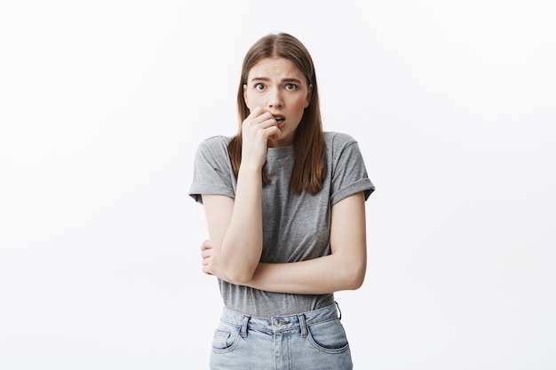 Portrait of unhappy beautiful young european student girl with dark hair and brown eyes in fashionable clothes gnaws fingers,  with scared expression, worried about she got pregnant.