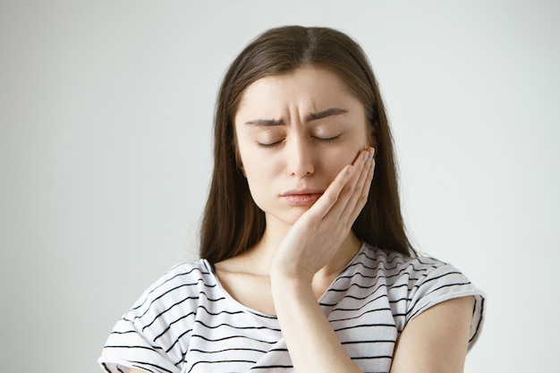 Free photo portrait of unhappy depressed young caucasian brunette woman closing eyes in frustration and pressing hand against cheek, trying to soothe pain while suffering from toothache, having tooth decay