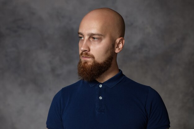 Portrait of unhappy grumpy bearded guy posing isolated . Upset sullen young Caucasian male with stubble and bald head havign pensive deep in thoughts facial expression. Human emotions