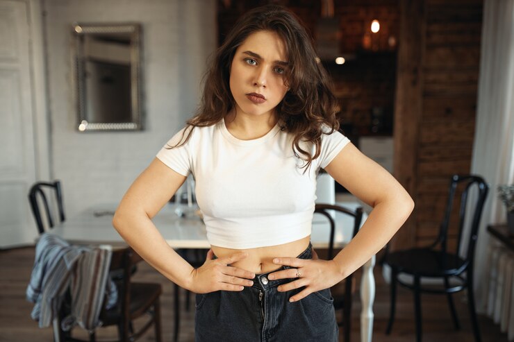 Portrait of unhappy grumpy young Caucasian woman in jeans and white top holding hands on her waist posing indoors