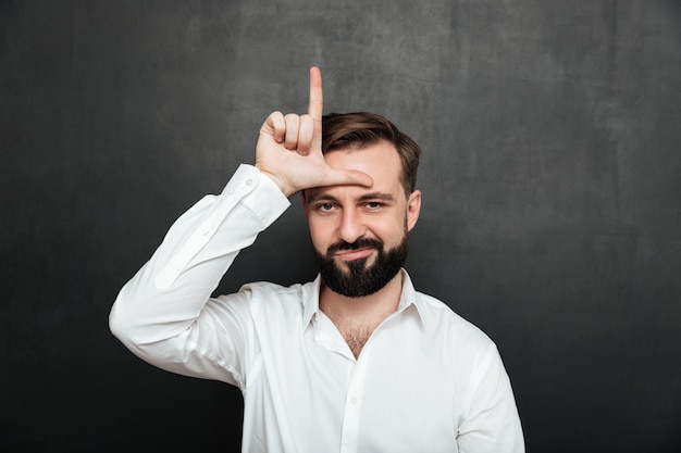 Free photo portrait of unhappy guy 30s posing on camera and showing loser sign on his forehead over graphite