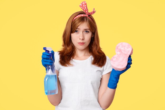 Portrait of unhappy housewife holds bottle with detergent and sponge, dressed casually, cleans house, poses on yellow. Displeased woman maid feels tired after spring cleaning