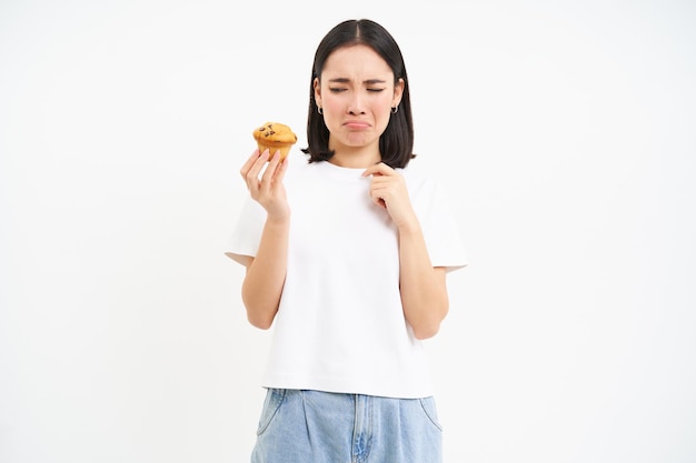 Foto gratuita il ritratto della donna coreana infelice guarda il bigné con la faccia triste ragazza sulla dieta non può mangiare il concetto di pasticceria
