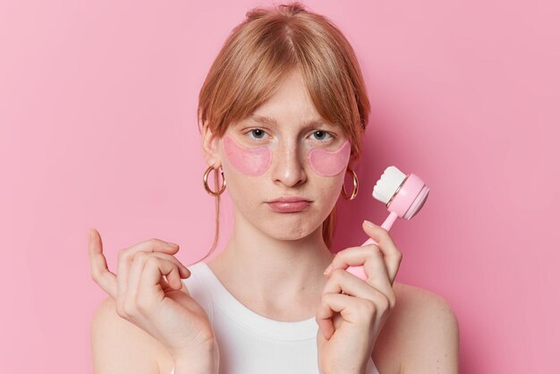 Portrait of unhappy redhaired girl has freckled skin applies beauty patches under eyes for skin treatment holds face massager dressed in casual white t shirt isolated over pink studio background