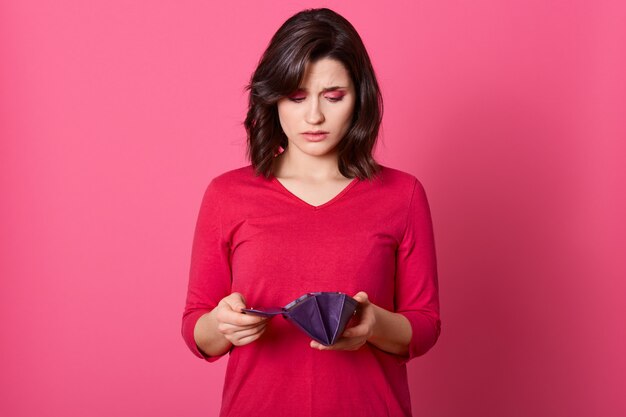 Portrait of unhappy stressed beautiful lady looking in open empty wallet with upset expression.