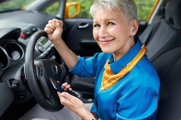 Portrait of unhappy stressed middle aged woman with shirt hairstyle sitting in driver's seat, clenching fist, holding mobile phone, dialing husband or calling roadside assistance because car is broken