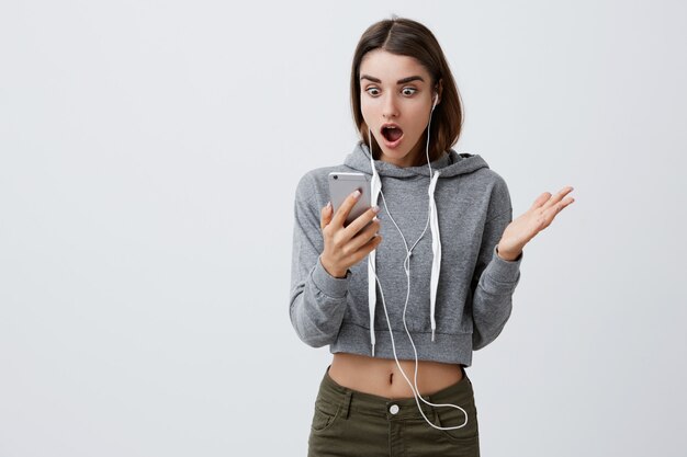 Portrait of unhappy young attractive caucasian student girl with long dark hair in casual stylish outfit looking in smartphone screen with shocked and angry expression, seeing photo of boyfriend